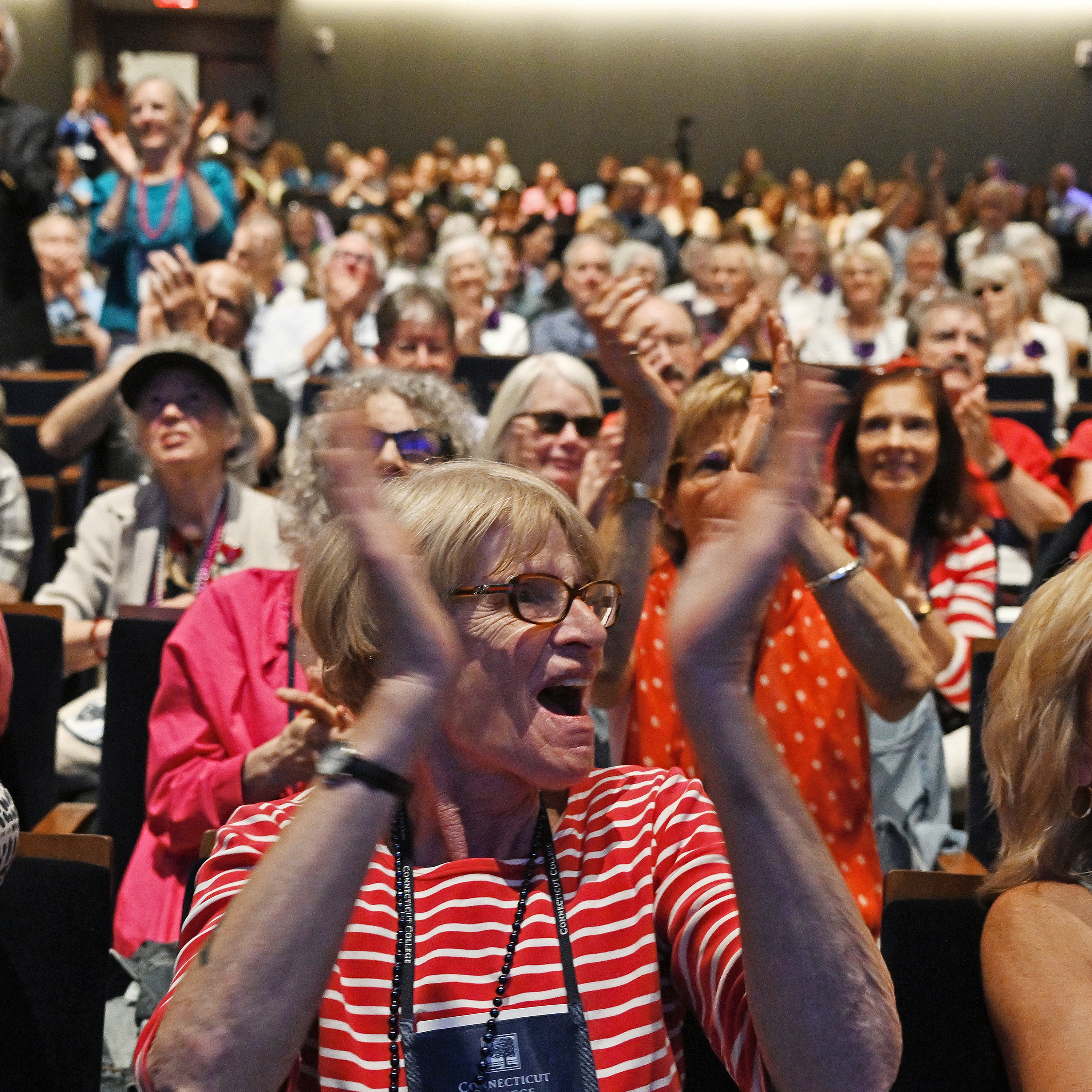 A crowd of college alumni gathered in a theater space applaud and cheer