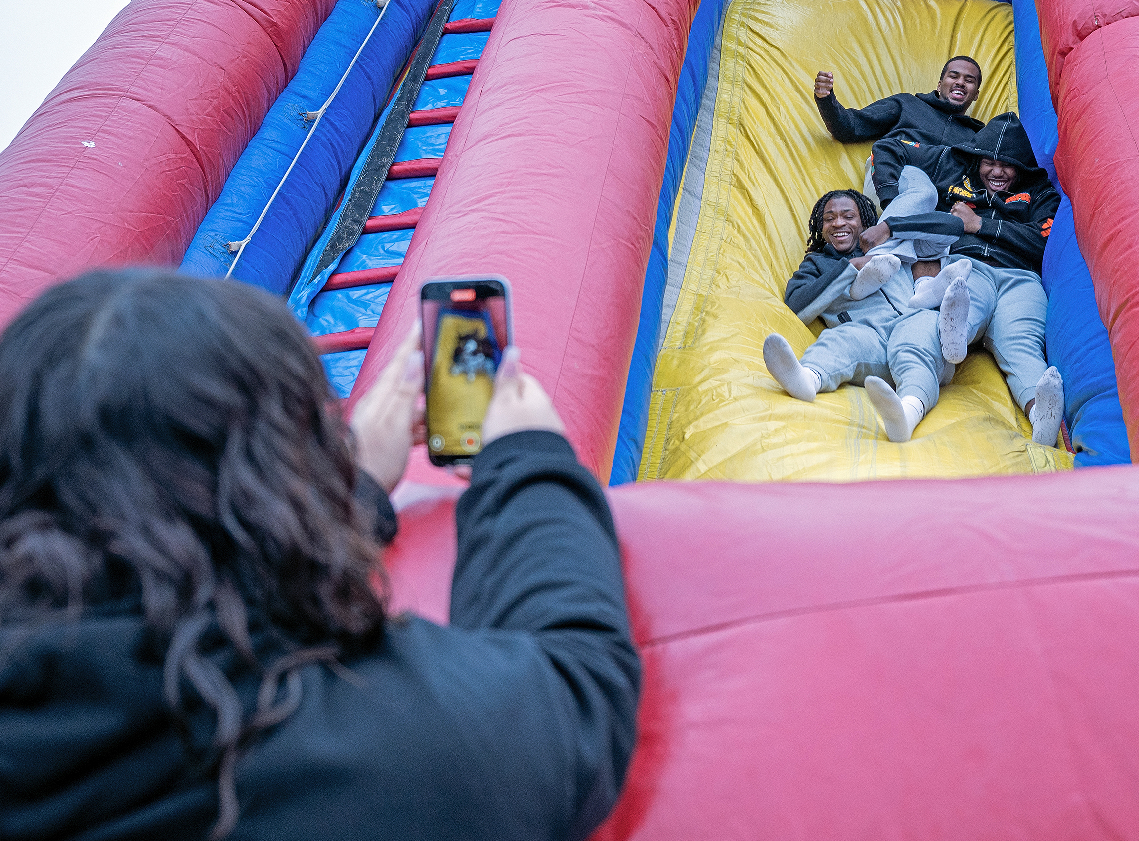 A student takes photos with a smart phone as three students ride down a giant inflatable bouncy slide.