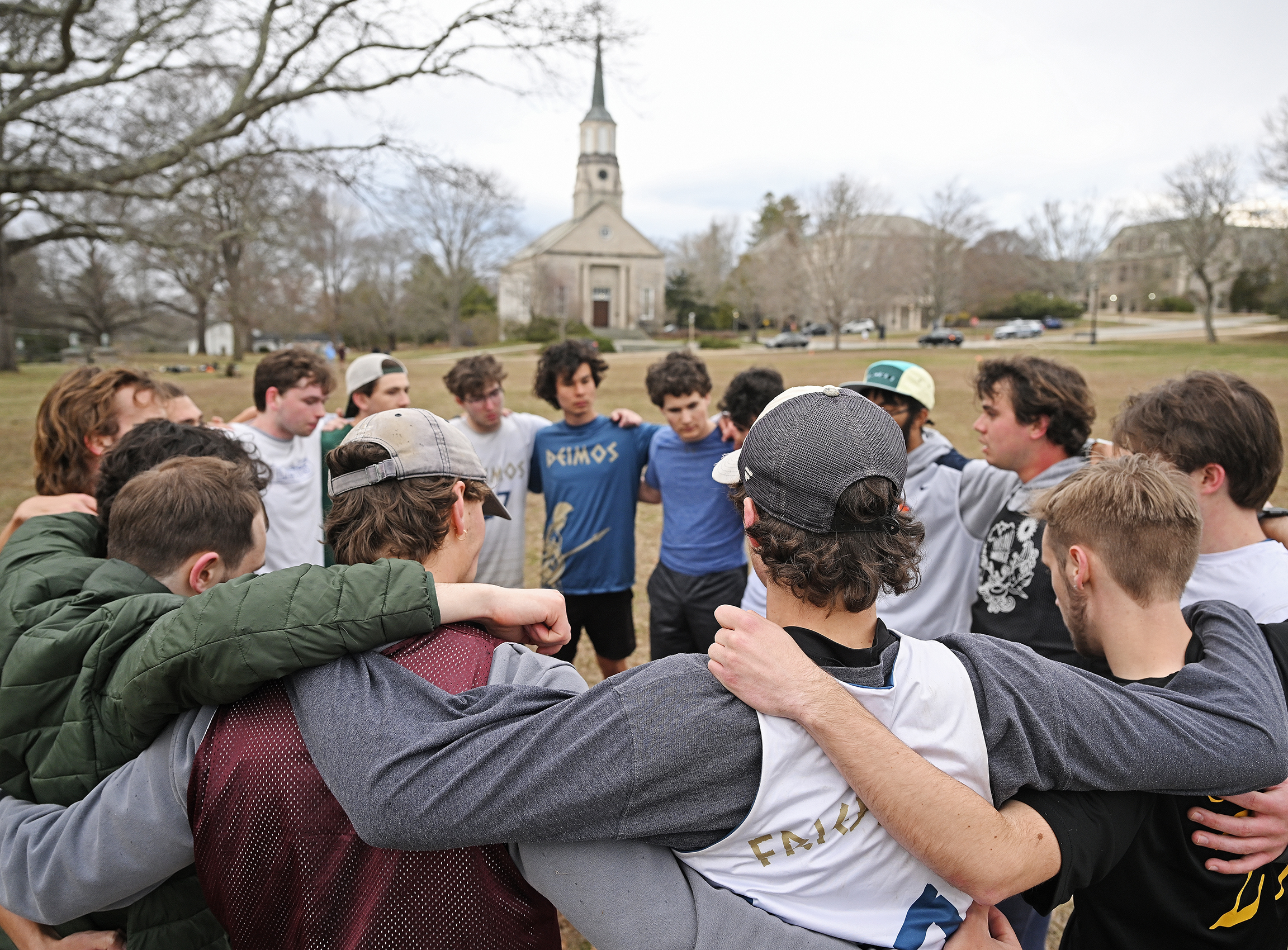 A group of student Ultimate Frisbee players huddle in a circle on a playing field with a church steeple visible in the distance.